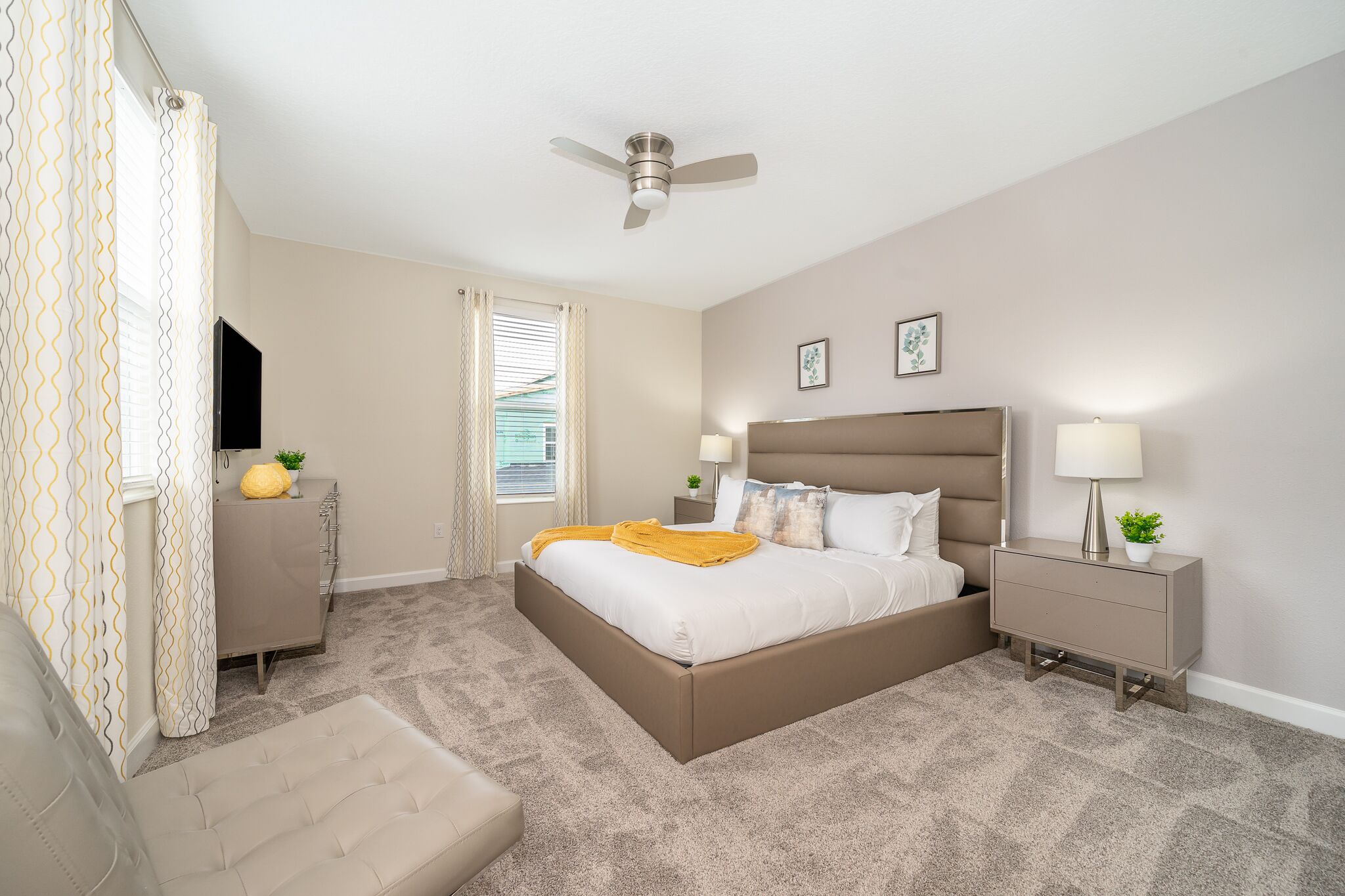 A bedroom with a bed, bedside tables, a ceiling fan, a window with curtains, and a wall-mounted TV.