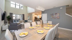 Ein moderner Essbereich mit einem langen Tisch, gedeckt für sechs Personen, einer Chandelieber und Blick in einen Wohnbereich mit einer Couch und einer Pflanze.