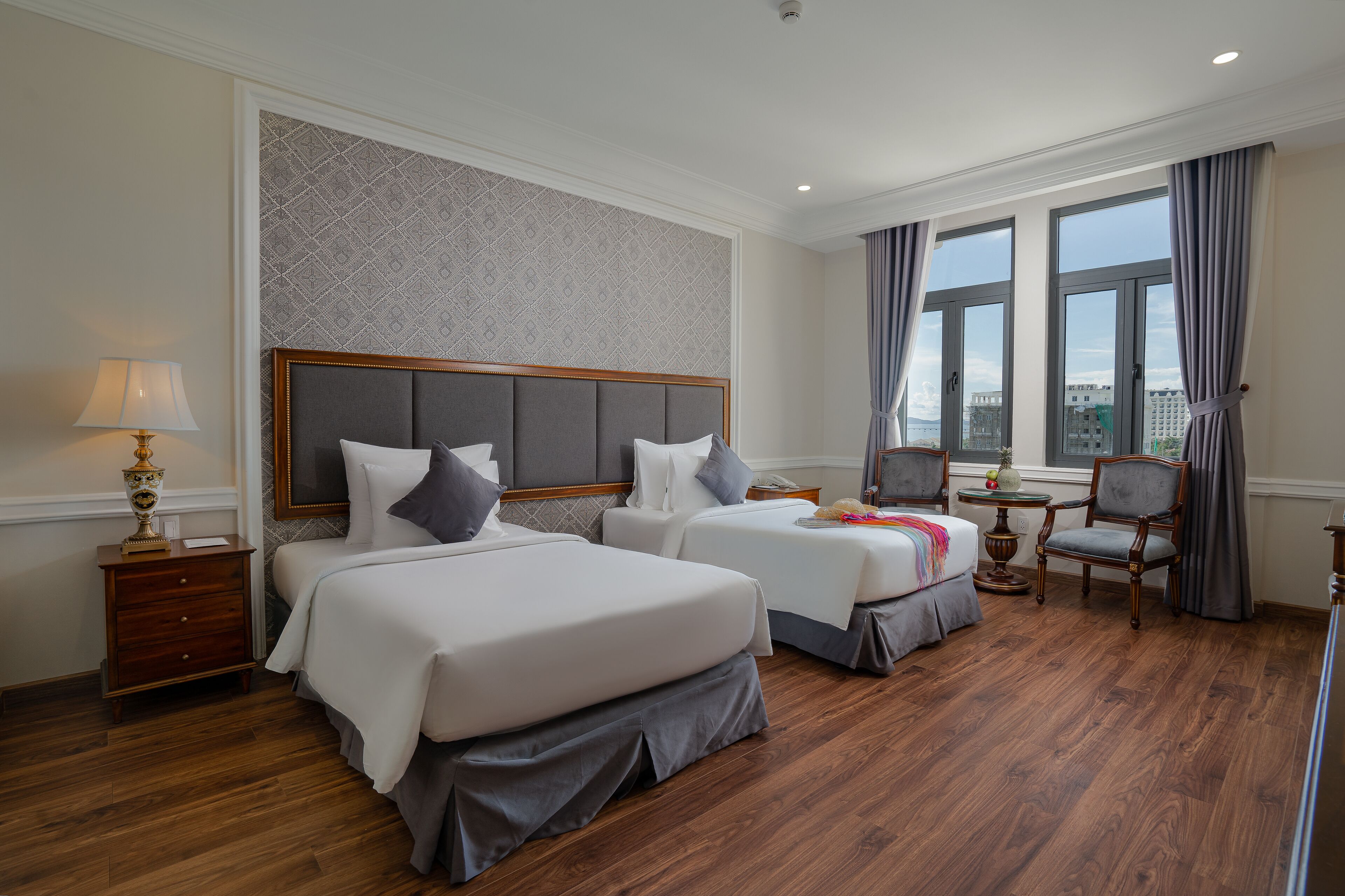 A hotel room with two beds, a small table, a chair, and a window with curtains.
