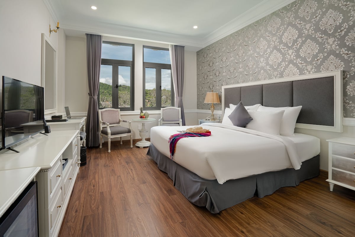 A hotel room with a large bed, a desk with a TV, a chair, and a view of the outdoors through large windows.