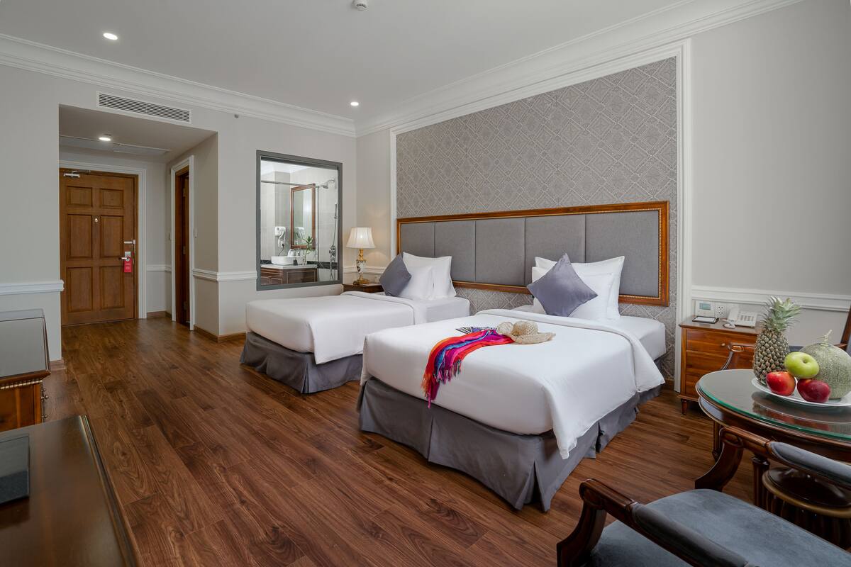 A hotel room with two beds, a wooden floor, a mirror, a small table with fruit, and a desk with a lamp.