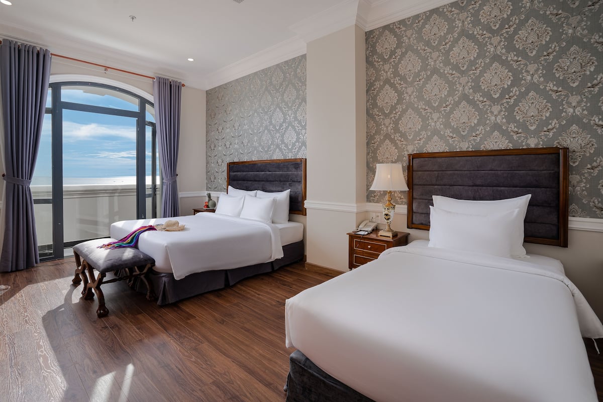 A hotel room with two beds, a large window with curtains, a wooden floor, and a patterned wallpaper.