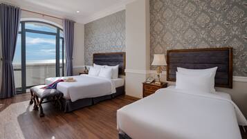 A hotel room with two beds, a large window with curtains, a wooden floor, and a patterned wallpaper.