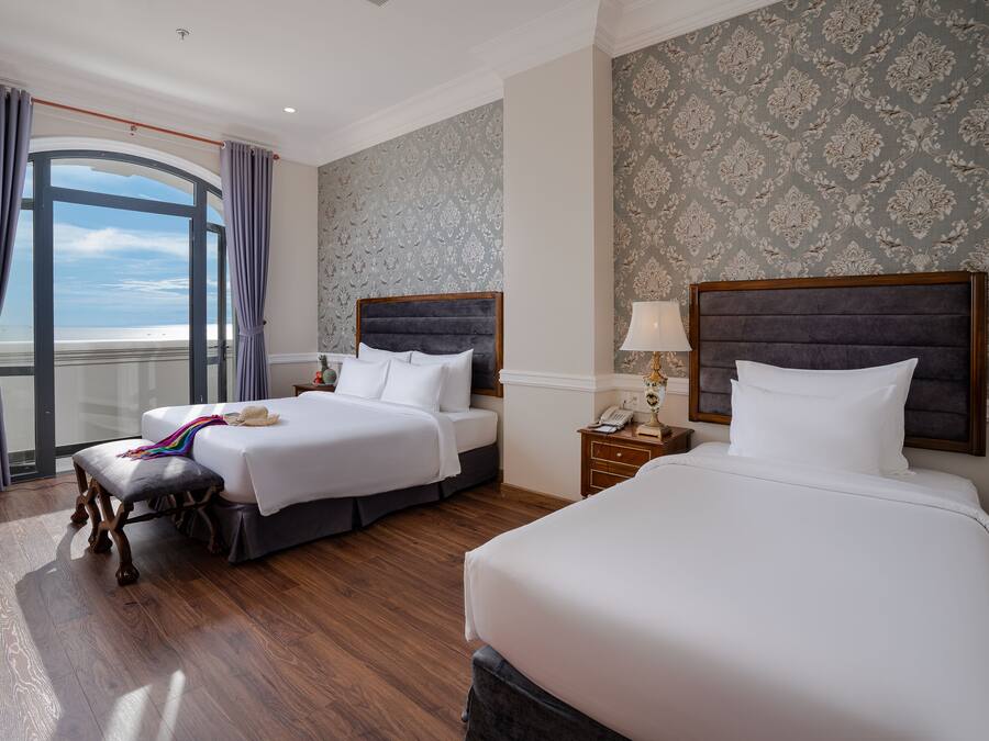 A hotel room with two beds, a large window with curtains, a wooden floor, and a patterned wallpaper.