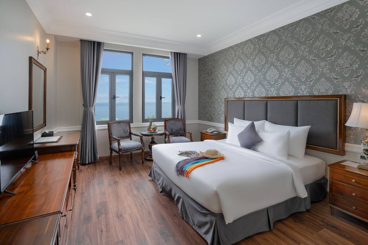 A hotel room with a large bed, a desk, a TV, and a view of the sea.