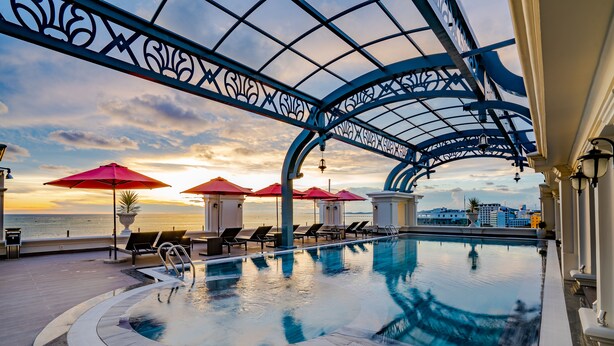 A rooftop infinity pool with lounge chairs and umbrellas, overlooking a cityscape and the sea.