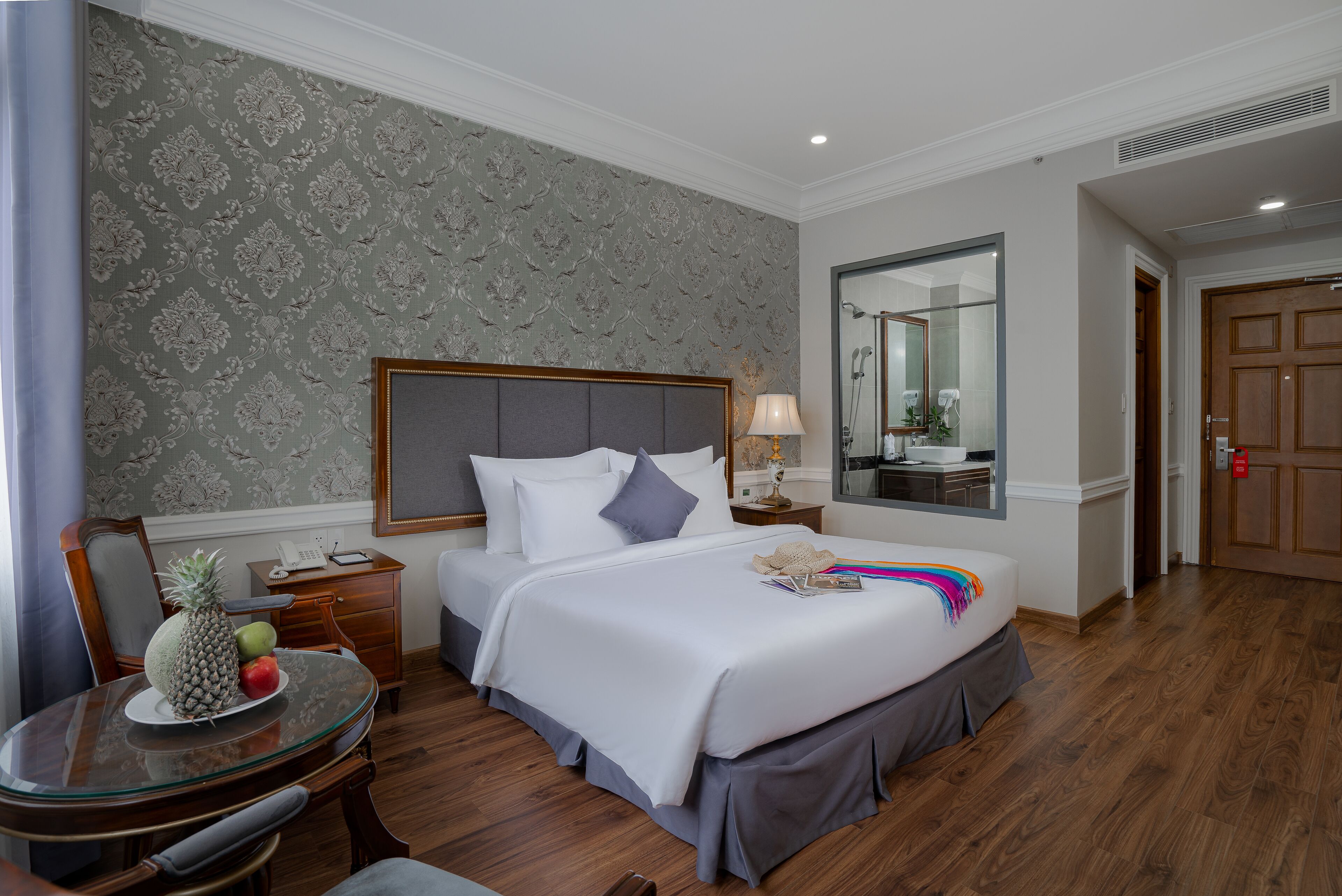 Habitación de hotel con una cama grande, una mesita de noche con lámpara, un espejo y piso de madera.