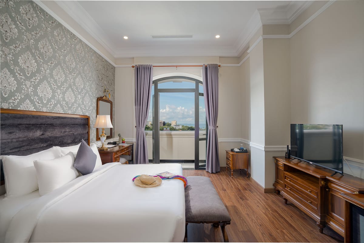 A hotel room with a large bed, a TV, a wooden dresser, and a balcony with curtains.