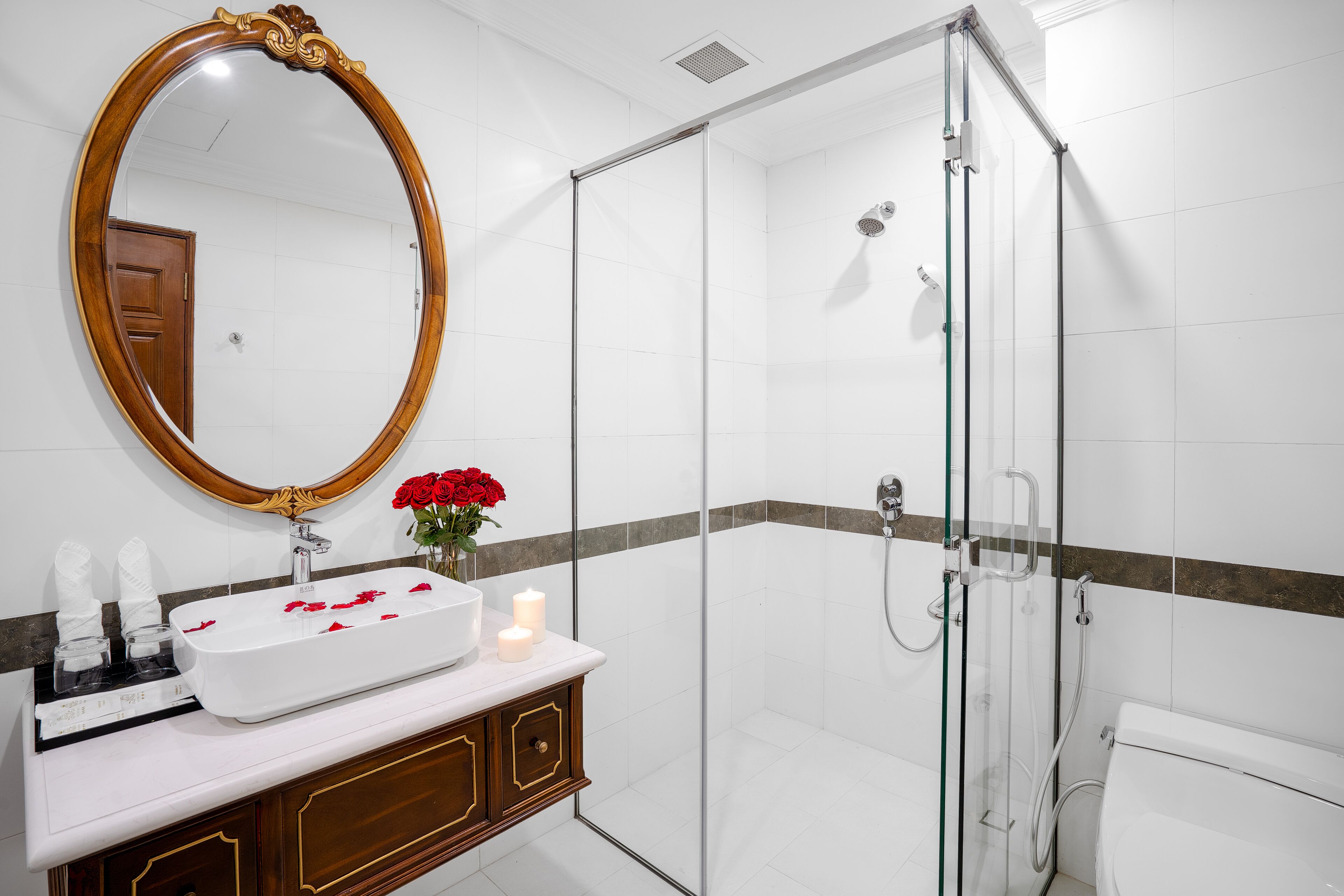A modern bathroom with a glass-enclosed shower, a large oval mirror, and a white sink with rose petals.