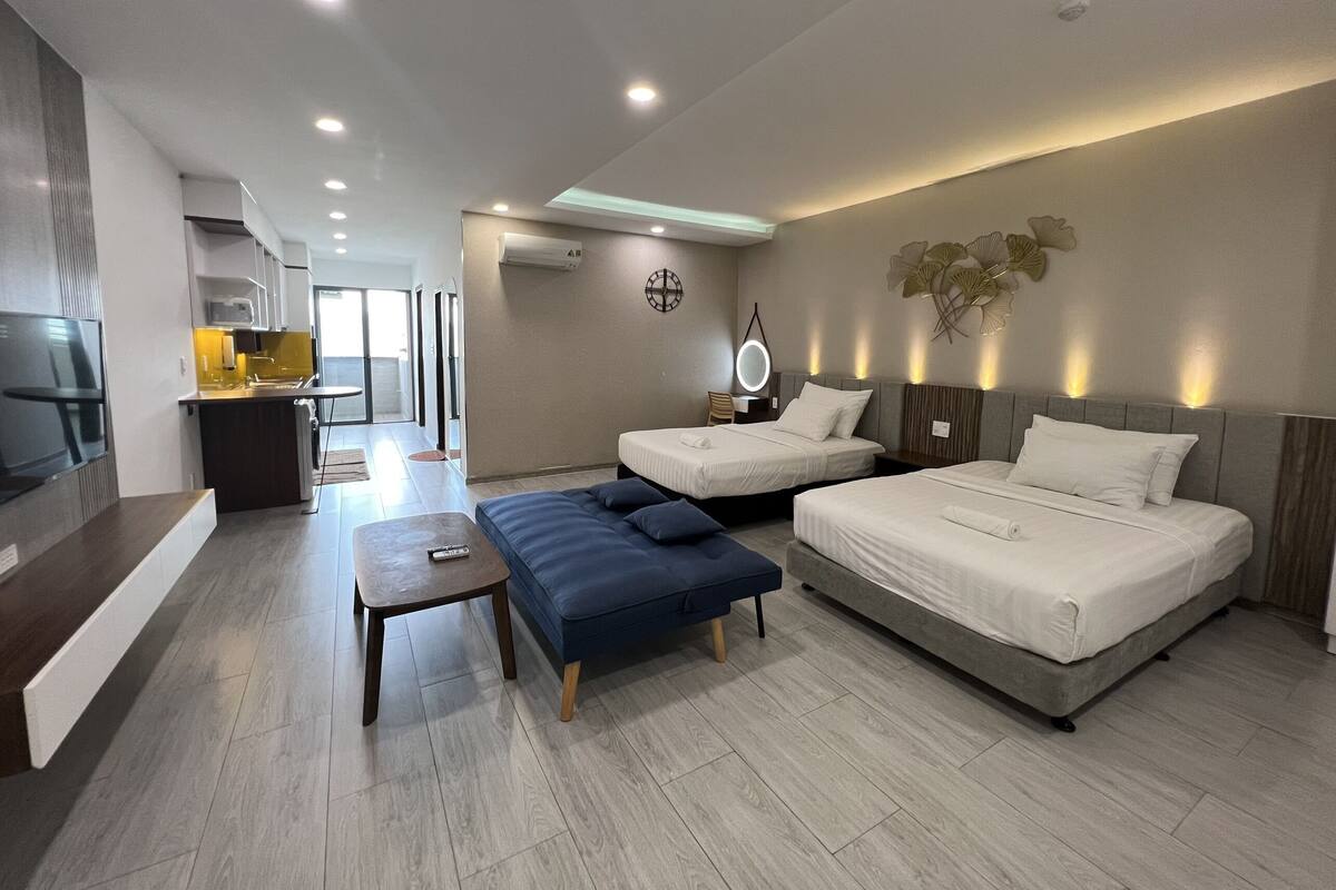 A modern hotel room with two beds, a sofa, a small table, a TV, and a kitchen area with a view.