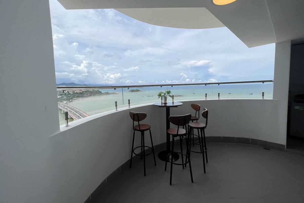 A balcony with a table and chairs, overlooking a coastal area with a bridge and boats.