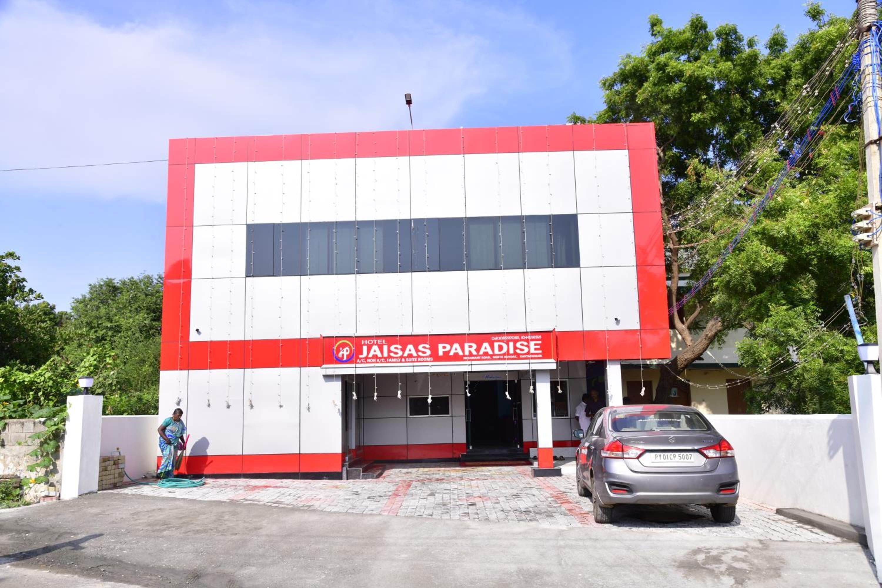 A hotel named "Jaisas Paradise" with a red and white facade, a car parked in front, and a person standing near the entrance.