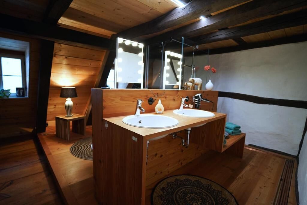 A wooden cabin bathroom with two sinks, a vanity, a mirror with lights, a small table with a lamp, and a rug on the floor.