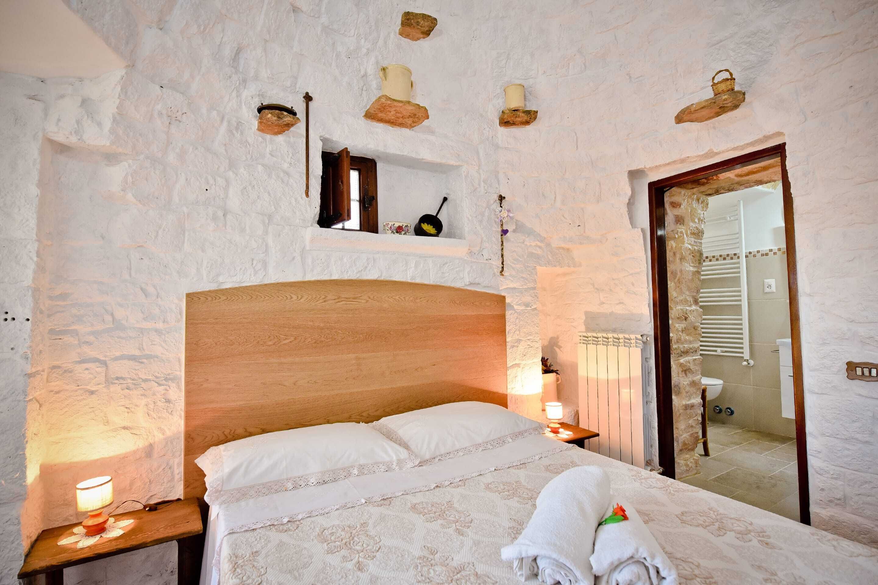 Quarto com cabeceira de madeira, mesa de cabeceira com abajur, prateleira pequena com objetos decorativos e vista para o banheiro por uma porta aberta.