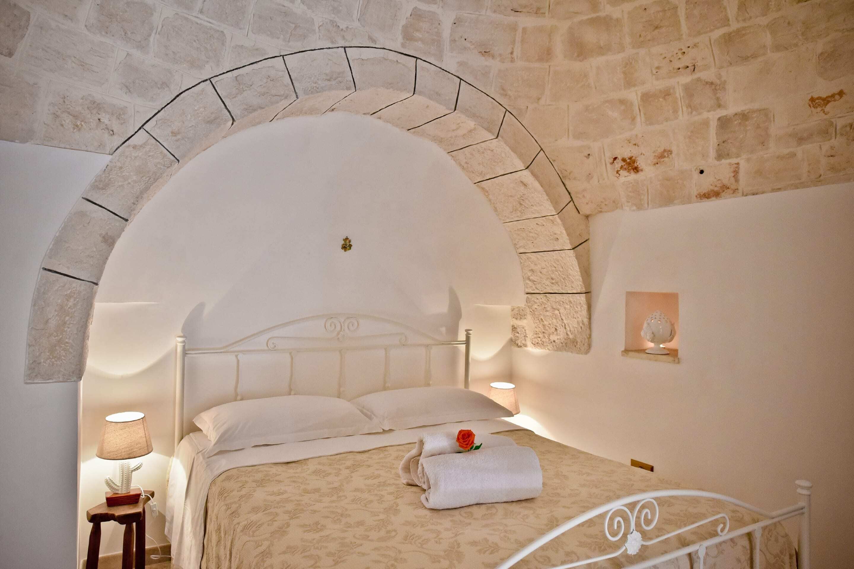 A bedroom with a stone wall, a bed with a white headboard, a nightstand with a lamp, and a small shelf with a decorative item.