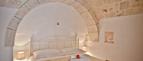 A bedroom with a stone wall, a bed with a white headboard, a nightstand with a lamp, and a small shelf with a decorative item.