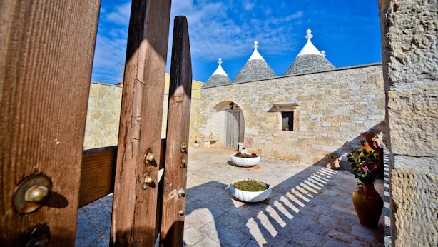 A courtyard with stone walls, a wooden door, and a view of a building with conical roofs. - I Cinque Trulli with Swimming Pool (Martina Franca)