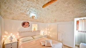 A bedroom with a stone ceiling, a bed with white bedding, a bedside table with a lamp, and a wall-mounted shelf with decorative items. - I Cinque Trulli with Swimming Pool (Martina Franca)