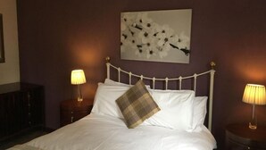 Ein ordentlich bezogenes Bett mit weißen Leinen und einem Karokissen, begleitet von zwei Nachttischlampen auf passenden Nachttischen. Über dem Bett hängt ein großes Blumenbild an der Wand.