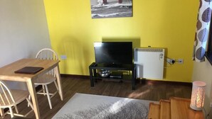 Ein Zimmer mit gelber Wand, einem Holztisch mit Stühlen, einem Fernseher auf einem Schrank, einem Heizkörper und einer Lampe.