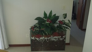 A potted plant with red flowers and green leaves in a corner of a room with white walls and a tiled floor. - Suitable Season Rio das Ostras 021991484253 (Rio das Ostras)