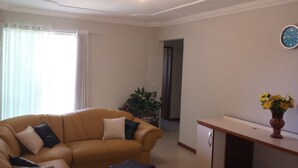 A living room with a beige sofa, a small table, and a potted plant. - Suitable Season Rio das Ostras 021991484253 (Rio das Ostras)
