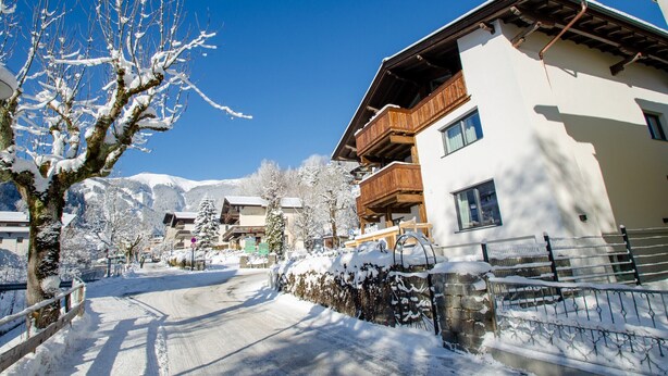 A snow-covered street with buildings, including a hotel with balconies and a wooden fence. - Finest Villa Zell am See - Skihaserl (Zell am See)