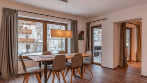 A dining area with a wooden table and chairs, large windows with curtains, and a hanging light fixture. - Finest Villa Zell am See - Skihaserl (Zell am See)