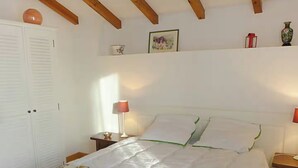 Un dormitorio con cama, paredes blancas, vigas de madera, una mesita de noche, una lámpara y una estantería con adornos.
