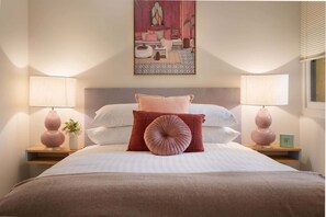 A bed with a headboard, two bedside tables with lamps, a wall-mounted painting, and a window with blinds. - Cosy 2-Bed Apartment in the Heart of Strathfield (Strathfield)