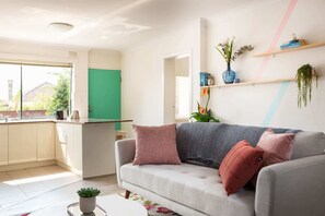 A modern living room with a grey sofa, a kitchen area with a green door, and a window with a view of a house. - Cosy 2-Bed Apartment in the Heart of Strathfield (Strathfield)