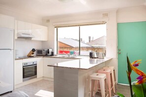 A modern kitchen with white cabinets, a built-in oven, a microwave, and a coffee maker. There is a window with a view of a building outside. - Cosy 2-Bed Apartment in the Heart of Strathfield (Strathfield)