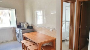 Pieni keittiö, jossa on pyykinpesukone, puinen pöytä penkeillä ja avoimen oven kautta näkyvä näkymä makuuhuoneeseen.