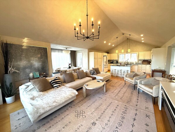 A spacious living area with a sofa, coffee table, and chairs, a kitchen with white cabinets, and a dining area with stools.