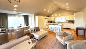 Ein modernes Wohnzimmer mit einer Couch, Sesseln, einem Couchtisch und einem Essbereich mit Tisch und Stühlen. Die Küche verfügt über weiße Schränke, einen Kühlschrank und eine Bar mit Hockern.