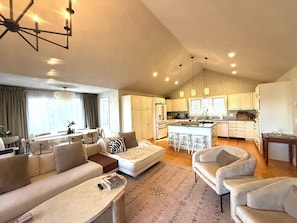 A modern living room with a sofa, armchairs, a coffee table, and a dining area with a table and chairs. The kitchen features white cabinets, a refrigerator, and a bar area with stools.
