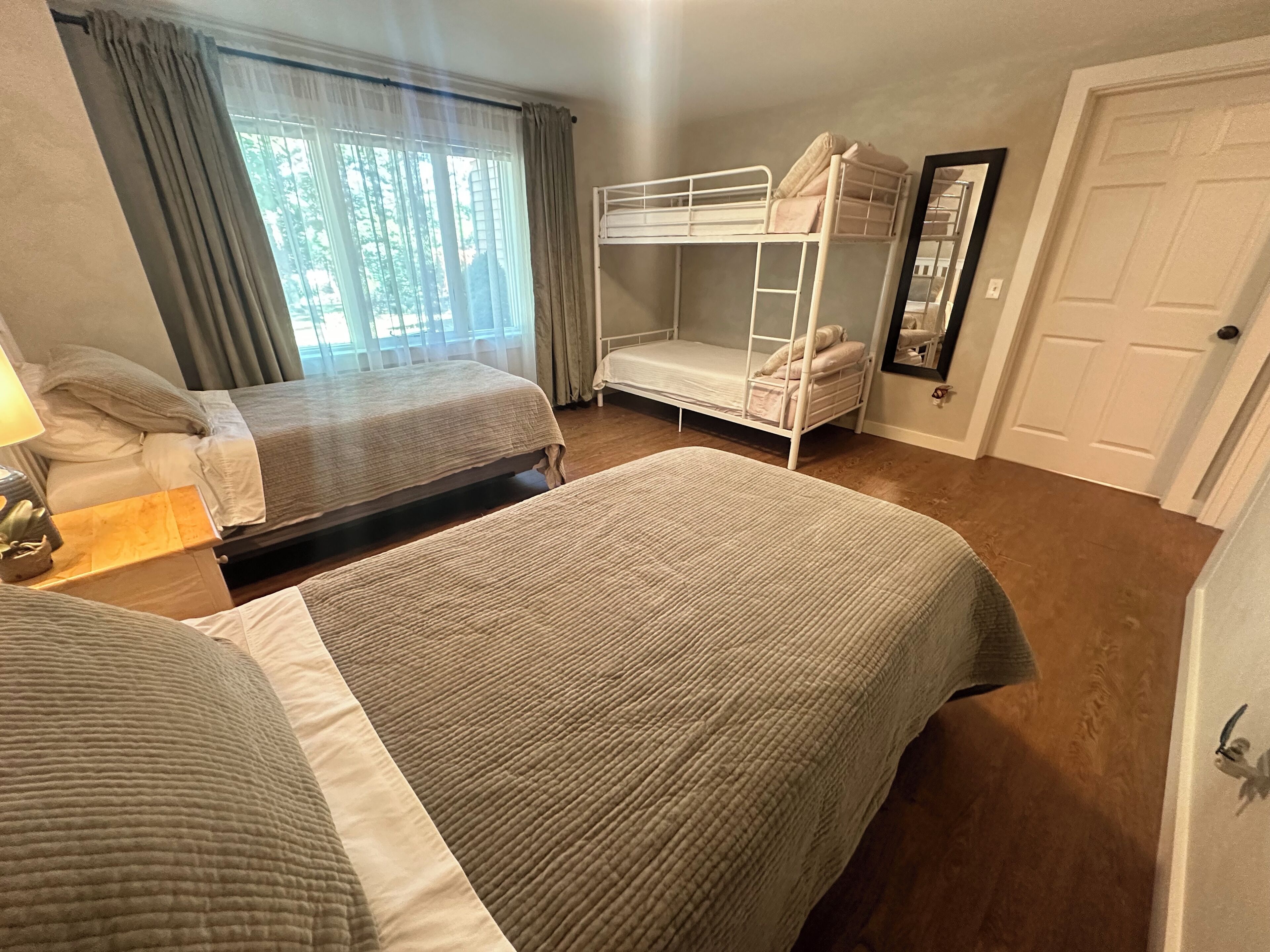 A room with two bunk beds, a bedside table with a lamp, a window with curtains, and a door.