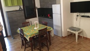 Een kleine keuken met een eettafel, stoelen, een koelkast, een televisie en een kastje.