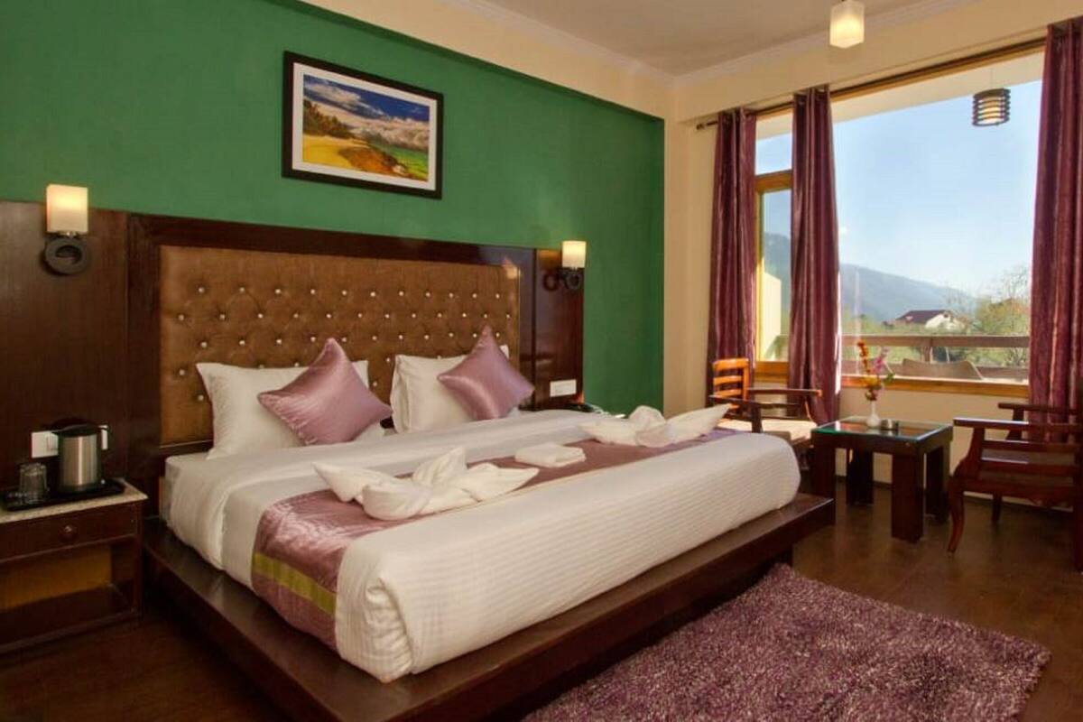A hotel room with a large bed, a wooden headboard, a green wall, a painting, a window with curtains, and a view of the outdoors.