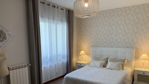 Ein Schlafzimmer mit einem großen Bett, zwei Nachttischen mit Lampen, einem Heizkörper und einem Fenster mit Vorhängen.