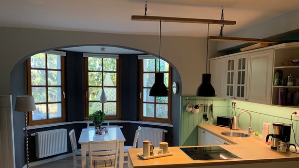 A kitchen with a central island, white cabinetry, a window with a view, and a hanging light fixture. - Ferienwohnung Seerose (Bleckede)