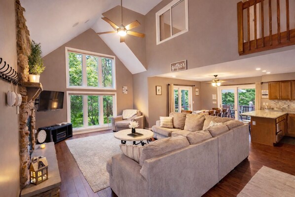 A spacious living area with a large sofa, a fireplace, and a television. There is a dining area with a table and chairs, and a kitchen with wooden cabinets.