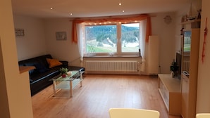 Una sala de estar moderna con un sofá negro, una mesa de centro de vidrio y vistas a un paisaje verde a través de amplios ventanales.