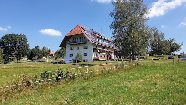 Un edificio de dos pisos con techo de tejas rojas y paneles solares, rodeado de un campo cubierto de hierba y árboles.