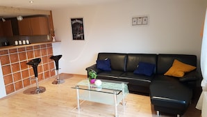 Una sala de estar con un sofá de cuero negro, una mesa de centro de vidrio y una cocina con armarios de madera y salpicadero de azulejos.