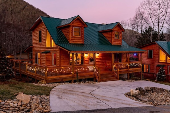 A log cabin with a green roof, a porch with string lights, and a Christmas tree.