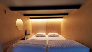 A compact hotel room with a bed, two pillows, a bedside table with a lamp, and a wall-mounted circular light fixture. - Green Marmot Capsule Hotel Zurich (Zürich)