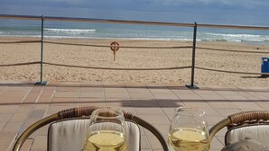 Vista de la playa con barandilla, un salvavidas y dos copas de vino blanco sobre una mesa.