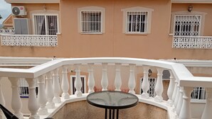 Un balcón con una mesa redonda y dos sillas, un edificio con techo de tejas rojas y ventanas con rejas.