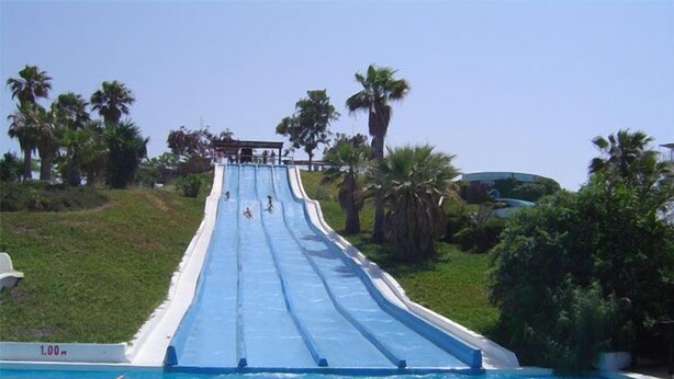 Una piscina con tobogán de agua, rodeada de palmeras y un cielo despejado.